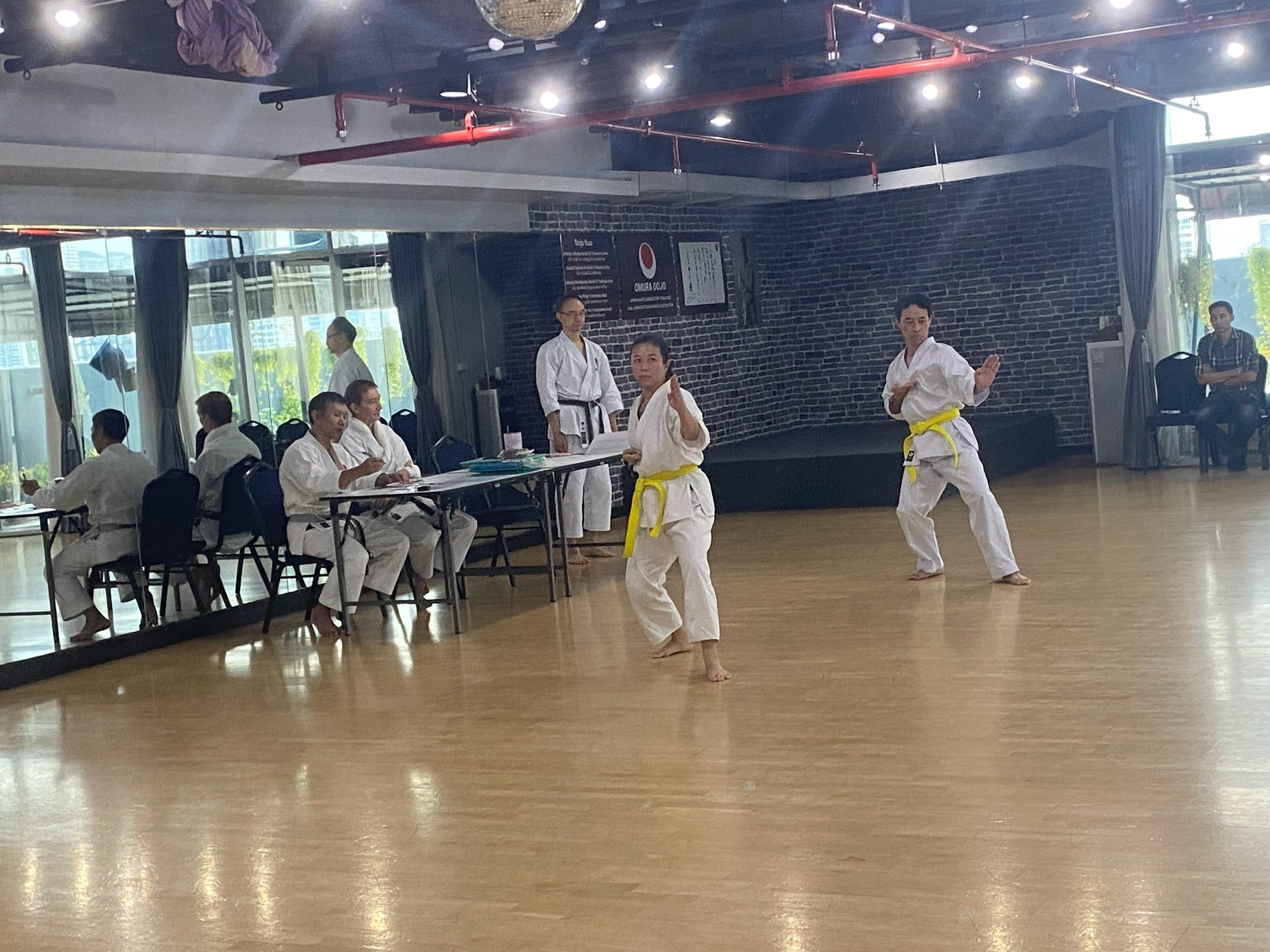 Two karateka in yellow belts performing kata at a grading, with examiners seated at a table